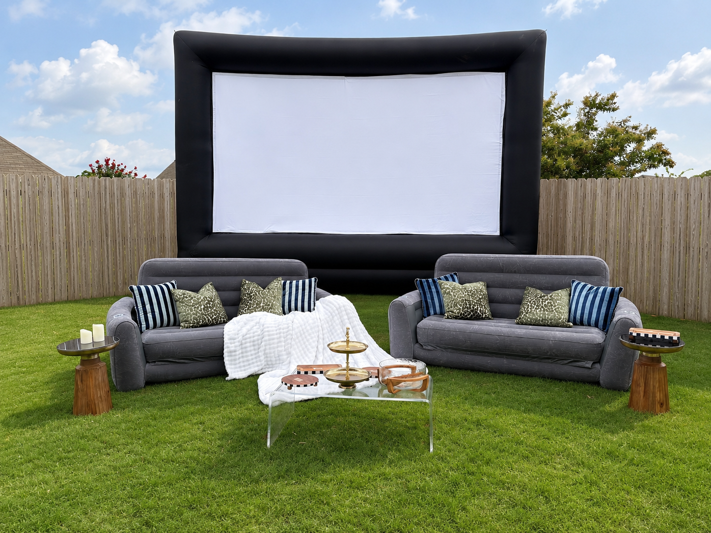 The Speakeasy setup with two grey inflatable couches facing a large inflatable screen, styled pillows, a blanket, and a glass coffee table