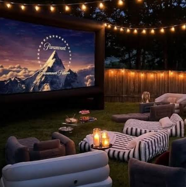 Backyard outdoor cinema at dusk with a large inflatable screen, string lights, candles, and striped inflatable lounge seating
